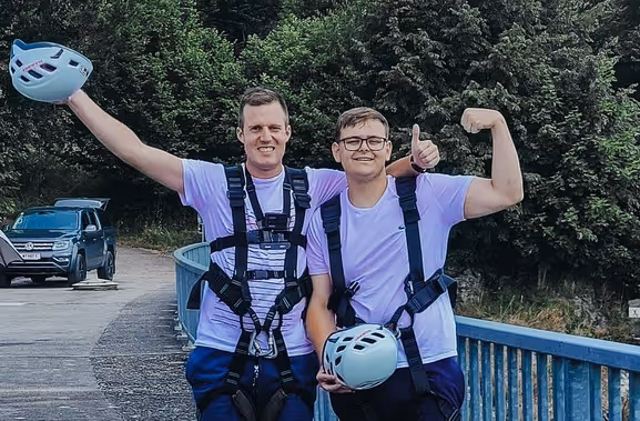 Zwei junge Männer stehen auf einer Brücke im Freien und tragen Sicherheitsgurte sowie Helme in der Hand. Sie lächeln in die Kamera und posieren selbstbewusst.
