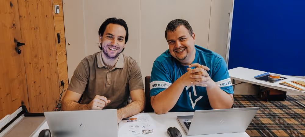 Zwei Männer sitzen nebeneinander an einem Tisch mit aufgeklappten Laptops und lächeln in die Kamera. Vor ihnen liegen Unterlagen und Arbeitsmaterialien.