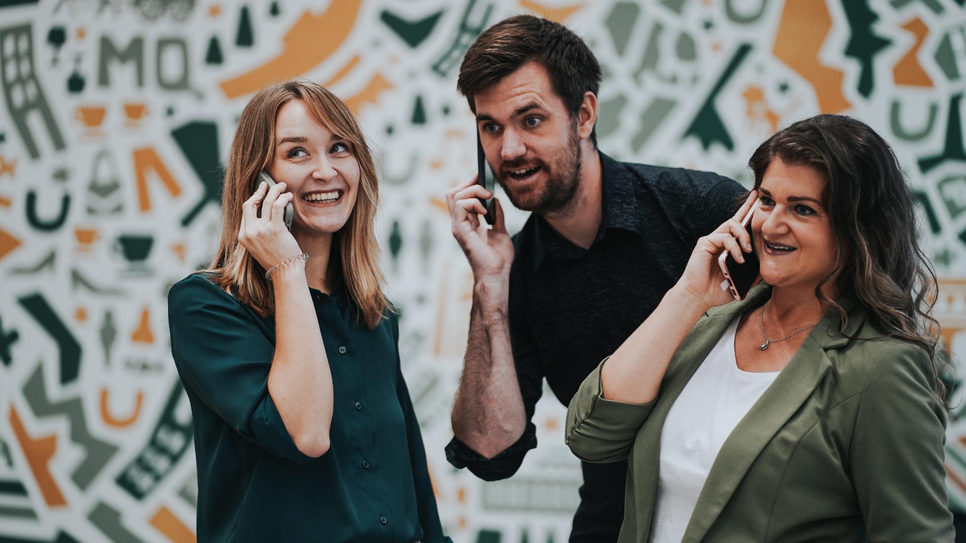 Cornelia Behrends und zwei Mitarbeiter lachen und halten ein Mobiltelefon an ihr Ohr.