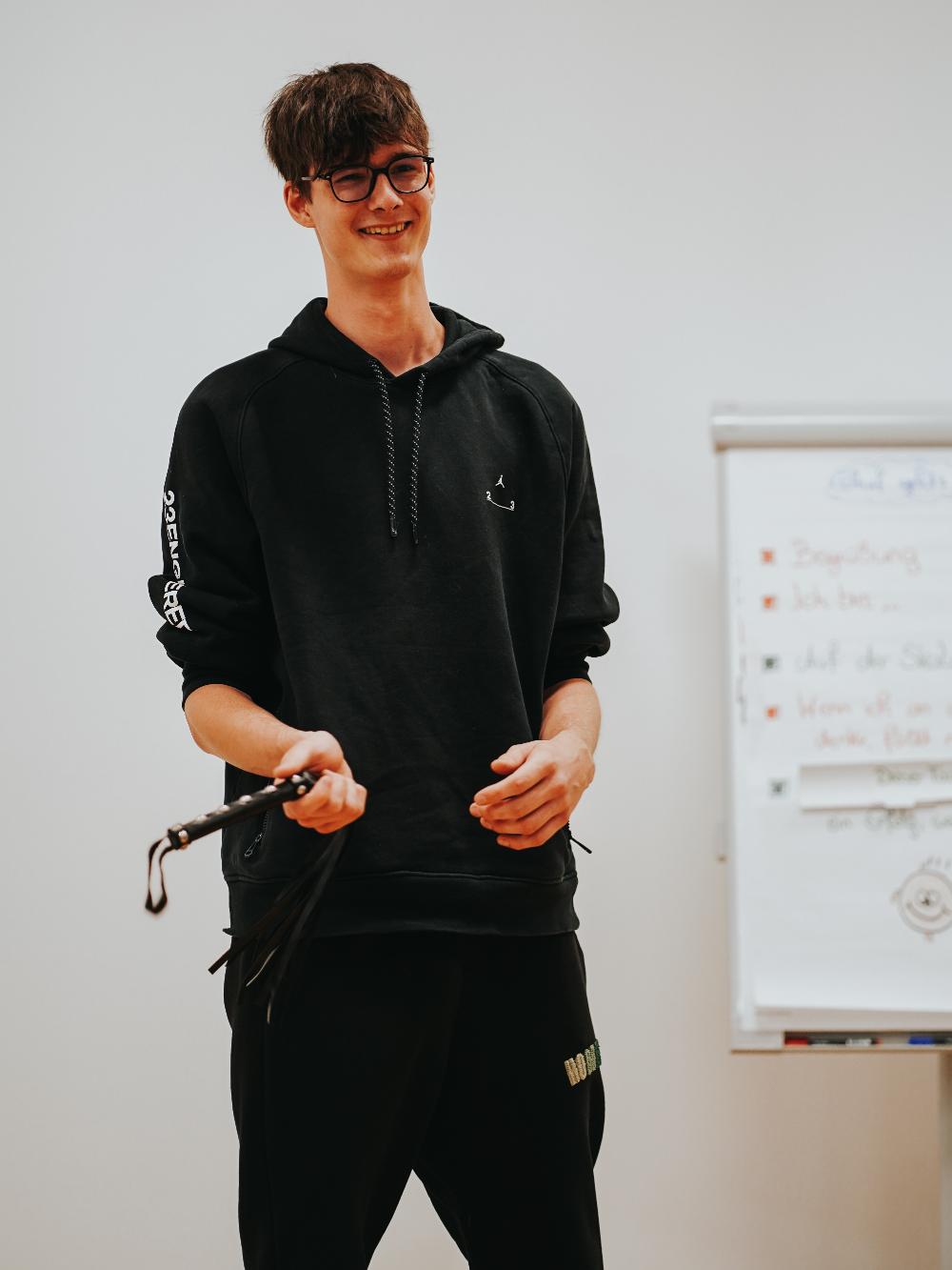 Ein junger Mann mit Brille, schwarz gekleidet, hält einen Gegenstand in der rechten Hand.