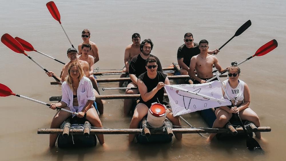 Eine Gruppe junger Menschen, welche auf einem selbstgebauten Floss im Wasser sitzen.