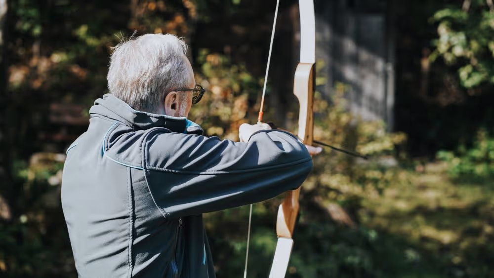 Ein älterer Mann mit grauem Haar und Brille steht im Freien in einem bewaldeten Gebiet und spannt einen Bogen, um einen Pfeil abzuschießen.